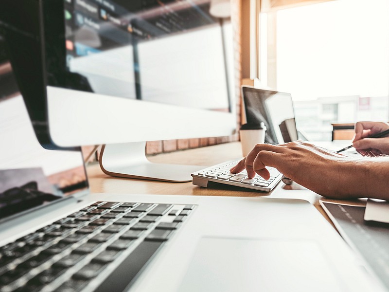 Person arbeitet am Schreibtisch mit Computer und Laptop, Hand tippt auf Tastatur, symbolisiert modernes Arbeiten und Technologie.